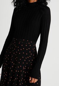 Schwarzer gerippter Rollkragenpullover mit langen Ärmeln, kombiniert mit einem schwarzen Rock mit braunen Punkten, der ein kontrastierendes Muster erzeugt.