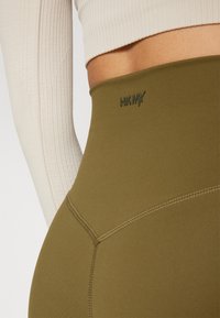 High-waisted olive green leggings in a stretchy fabric, featuring a fitted silhouette and contrasting stitching with a subtle logo.