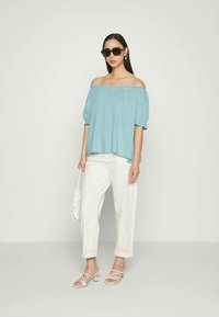 Mulher vestindo uma blusa azul clara de ombro à mostra, calças brancas com as barras arregaçadas, saltos brancos com tiras, segurando uma bolsa branca e óculos de sol pretos.