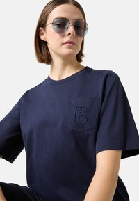 Námořnická modrá bavlněná tričko s kulatým výstřihem, krátkými rukávy a přední kapsou s vyšitým písmenem "B". Model má na sobě tmavé sluneční brýle.
