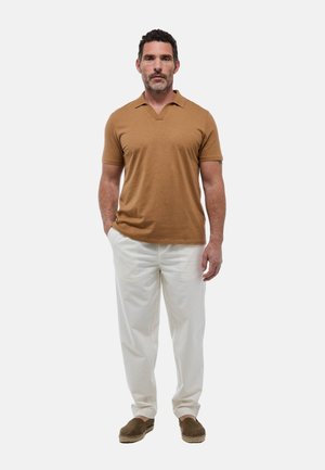 Homme debout les mains dans les poches, portant une chemise marron à manches courtes, un pantalon blanc et des chaussures marron sans lacets sur un fond uni.