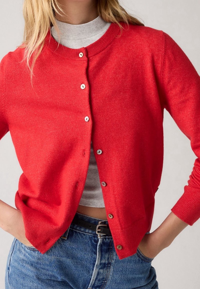 Femme portant un cardigan rouge vif boutonné sur une chemise grise claire avec un jean bleu, les mains dans les poches, cadrée au niveau du cou et des hanches.