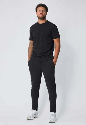 Homme aux cheveux courts et bouclés avec des tatouages, portant un t-shirt noir et un pantalon avec des baskets blanches, se tenant contre un fond clair uni.