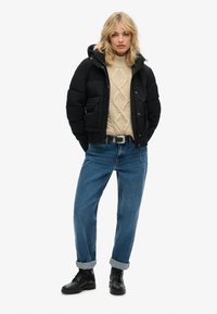 Zwarte gewatteerde jas met capuchon, beige kabelgebreide trui, blauwe opgerolde jeans en zwarte veterschoenen. Focus op een comfortabele en gelaagde outfit.