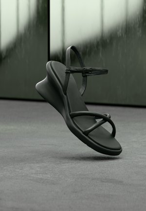 Juoda minimalistinė sandalė su plonais dirželiais ir sagties užsegimu, rodoma priešais neryškų žaliai pilką foną.