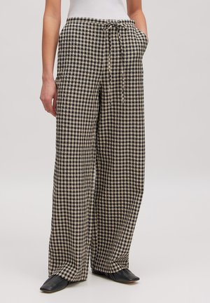 Femme portant un pantalon large à carreaux noir et beige avec une taille à cordon, associé à des chaussures pointues noires et un haut blanc.