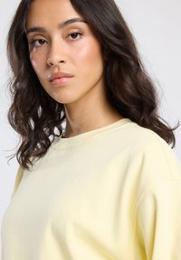 Young woman with wavy dark hair wearing a pale yellow crewneck shirt, looking slightly to the side against a light background.