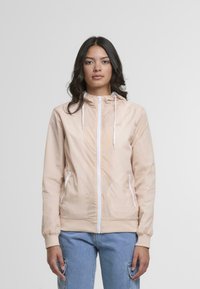 Ženska z darki lasmi, oblečena v bež jakno na zadrgo in svetlo modre hlače s šivi, stoji ob običajni svetlo sivi ozadju.