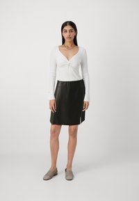 Camiseta blanca de manga larga con escote retorcido, combinada con una falda de cuero negra con una abertura lateral, complementada con zapatos de tela gris.