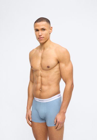 Μυώδης άνδρας με κοντά μαλλιά, ντυμένος με ανοιχτό μπλε μποξεράκια με λευκή ζώ νη, στέκεται μπροστά σε απλό λευκό φόντο.