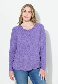 Fioletowa bluzka z długim rękawem, w kwiatowy wzór, uszyta z miękkiego materiału, z okrągłym dekoltem i lekko zaokrąglonym dołem. Dobrana z niebieskimi dżinsami.