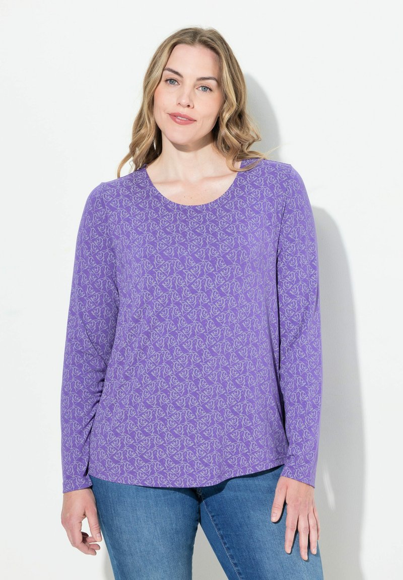 Fioletowa bluzka z długim rękawem, w kwiatowy wzór, uszyta z miękkiego materiału, z okrągłym dekoltem i lekko zaokrąglonym dołem. Dobrana z niebieskimi dżinsami.