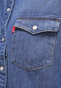 Chemise en denim avec une poche sur la poitrine à fermeture à pression, de couleur bleu clair, texture subtile et couture jaune contrastante. Étiquette de marque rouge visible.