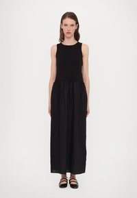 Robe maxi noire avec un haut sans manches, un corsage ajusté et une jupe froncée. Fabriquée dans un tissu léger au toucher doux. Chaussures plates à lanières.