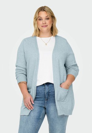 Femme aux cheveux blonds portant un cardigan ouvert bleu clair, une chemise blanche, un jean bleu et un collier en or, se tenant avec une main dans la poche.