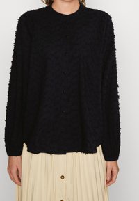 Svart, långärmad blus i texturerad tyg, rund hals och knappstängning. Mjuk, avslappnad passform med en något flödande silhuett.