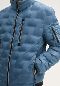 Padded jacket w jasnoniebiesnym nylonie z pikowanym wzorem, z wysokim kołnierzem, dwoma zapięciami z przodu i kieszenią na zamek błyskawiczny na rękawie.