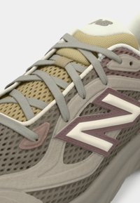 Gros plan sur une chaussure de sport New Balance beige et taupe en tissu mesh, avec des lacets gris croisés et le logo « N » sur le côté.