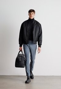 Schwarze Bomberjacke über einem schwarzen Rollkragenpullover, graue Hose und schwarze Stiefeletten. Trägt eine schwarze Tasche mit silbernen Akzenten.