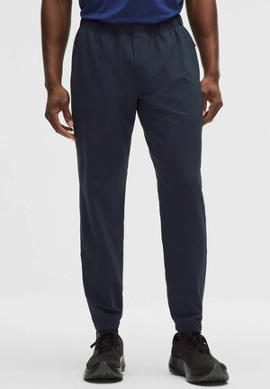 Hombre con pantalones jogger azul marino con cintura elástica y zapatos deportivos negros, de pie contra un fondo blanco.