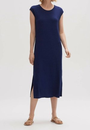 Mujer con vestido midi sin mangas azul marino con abertura lateral, combinada con sandalias planas beige, de pie frente a un fondo blanco liso.