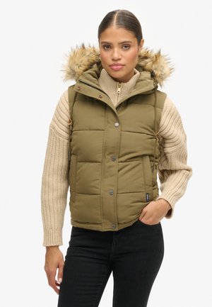 Gilet rembourré vert olive avec une capuche en fourrure amovible, des boutons-pression et deux poches latérales, porté sur un pull beige côtelé et un jean noir.