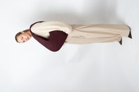 Femme portant un pantalon large beige et un pull crème, tenant un grand sac à bandoulière texturé de couleur bourgogne foncé, se tenant devant un fond blanc.