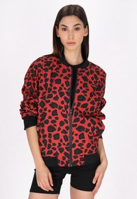 Giacca bomber rossa con stampa leopardata nera, caratterizzata da una chiusura a zip frontale, polsini a coste e dettagli neri. Indossata sopra una maglietta nera e pantaloncini.