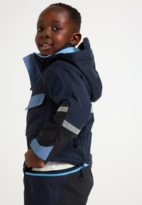 Giacca outdoor blu navy con accenti blu, dotata di strisce riflettenti, multiple tasche e cappuccio. Texture morbida e polsini regolabili.