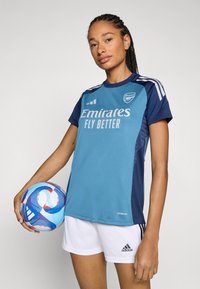 Turkos och marinblå fotbollsjersey med texten "Emirates Fly Better", i kombination med vita shorts. Flätat hår, håller en flerfärgad fotboll.