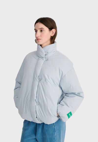 Femme portant une doudoune bleu clair avec un col haut et des boutons, les mains dans les poches, debout devant un fond clair uni.