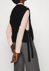 Gilet en tricot noir avec des côtés ouverts, orné d'une broche décorative à l'épaule. Porté sur une blouse rose clair et un pantalon gris à motifs.