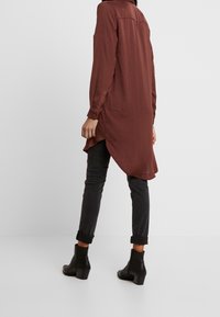 Personne portant un jean gris foncé retroussé, des bottines noires, et une longue chemise marron avec un ourlet arrondi, se tenant sur un sol et un fond blancs.