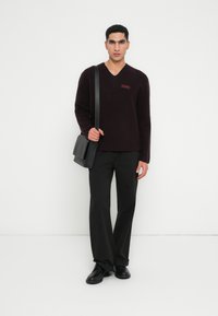 Βουργουνδί v-neck πουλόβερ με μακριά μανίκια, μια μικρή κόκκινη ετικέτα, συνδυασμένο με μαύρο παντελόνι και μαύρη τσάντα ώμου. Λείο υλικό.