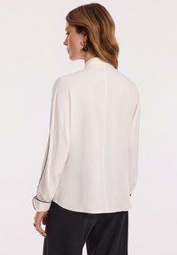 Biała bluzka o gładkiej, lekkiej fakturze, z długimi rękawami, wysokim kołnierzem i czarnym wykończeniem wzdłuż rękawów.