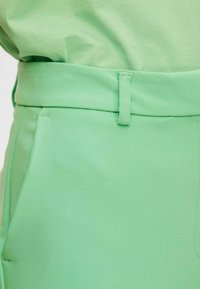 Pantaloni di stoffa verdi con una texture liscia, vita standard, tasche laterali e dettagli di cucitura precisi.