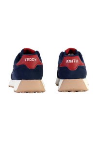 Scarpe sportive blu navy con dettagli rossi, accenti bianchi e suole beige. La parte posteriore presenta "TEDDY" e "SMITH" in lettere bianche.