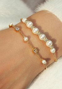 Tre braccialetti d'oro su un polso: uno con grandi perle e cristalli, uno con piccole perle alternate a accenti quadrati, e un altro con una gemma quadrata.