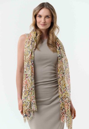 Frau mit welligem blondem Haar, die ein ärmelloses graues Kleid trägt und einen langen, mehrfarbig gemusterten Schal über die Schultern gelegt hat.