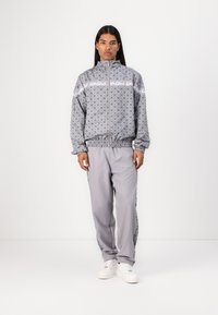 Grijze gepatterneerde trainingsoutfit met een ritsjack en bijpassende broek, elastische manchetten en logo-accenten. Draag met witte sneakers.