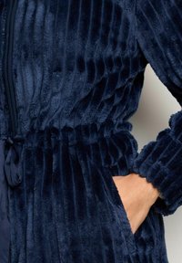 Accappatoio in pile blu navy con texture a righe, vita elastica e tasche. Tessuto liscio, finitura morbida e polsini arricciati per un maggiore comfort.