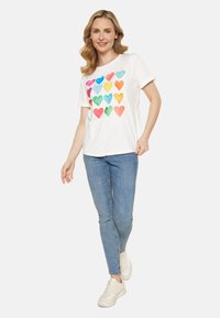 Hvid bomulds T-shirt med farverige hjerte mønstre i forskellige nuancer. Sammen med blå denim jeans og lyse sneakers.