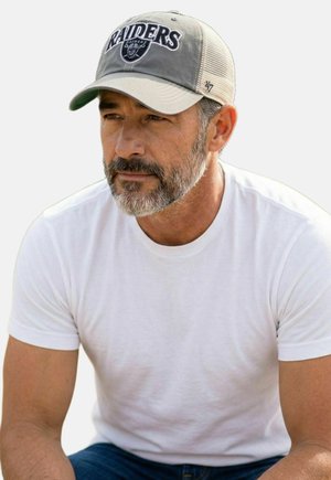 Middle-aged man with salt-and-pepper beard wearing Raiders baseball cap, white t-shirt, and jeans, looking focused and slightly to the side.