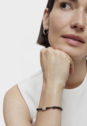 Czarna zaplatana bransoletka z szarym kwiatowym akcentem. Modelka w bezrękawniku, opierająca podbródek na dłoni. Prosty design, minimalna tekstura.