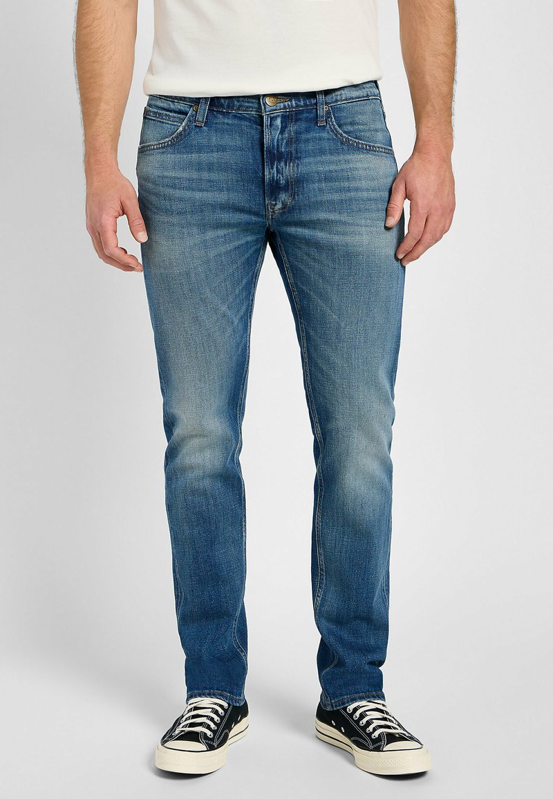 Jeans de corte slim para hombre en denim azul medio con desgastado y efecto de arrugas. Presenta cinco bolsillos y una cremallera con cierre de botón.