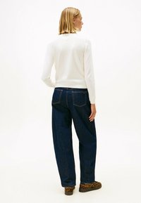 Frau mit blonden Haaren, die ein weißes Langarmoberteil, dunkelblaue weite Jeans und Schuhe im Leopardenmuster trägt, steht mit dem Rücken zu einem weißen Hintergrund.