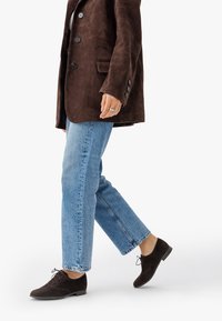 Giacca in suede marrone, jeans dritti blu chiaro e scarpe stringate marrone scuro. La giacca presenta bottoni grandi e una superficie liscia.