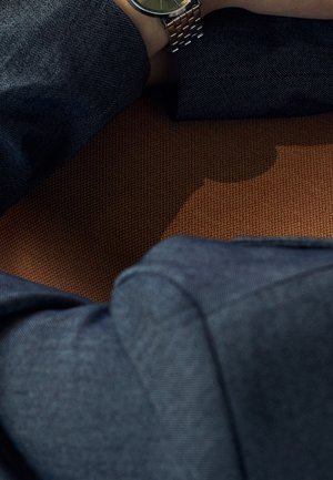 Close-up van gekruiste armen met een blauw shirt met lange mouwen en een zilveren polshorloge, rustend op een gestructureerd bruin oppervlak.