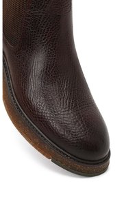 Botín de cuero marrón hasta el tobillo con un acabado texturizado, punta redondeada y suela de color marrón claro en contraste. Cuenta con paneles laterales elásticos para poner y quitar fácilmente.