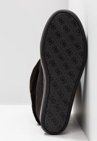 Suela de zapato negra con un patrón repetido de logotipo floral en relieve, inclinada contra un fondo blanco y una superficie vertical blanca.